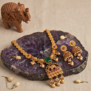 Traditional Heritage Style temple necklace set featuring an intricate elephant motif pendant, square green emerald stone, pink ruby accents, and matching jhumka earrings displayed on dark green velvet fabric.