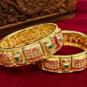 Pair of heavy gold-plated broad bangles featuring raised textured block panels, red ruby stone grid patterns, and square emerald green center stone accents from Glorious Jewels.