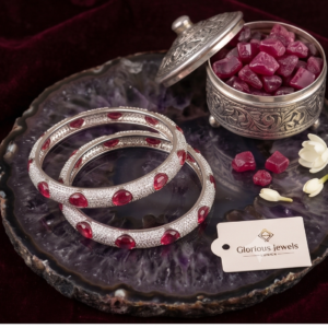 Pair of silver-plated micro-pave CZ bangles studded with oval ruby red gemstones resting on a purple agate slice next to an antique silver box filled with raw rubies.